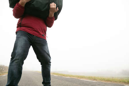 man in jeans and red mackintosh on foggy road, heavy suitcase on his back unrecognizable