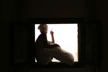 Woman with towel on her head, sitting in the window of a hotel room, smoking a cigarette, unrecognisable.の写真素材