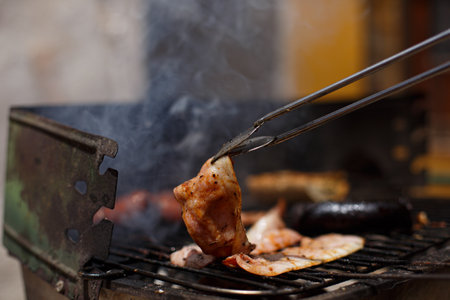 Hand with the help of metal tongs turns the simmering potatoes and sausages on a black barbecue grill.の写真素材