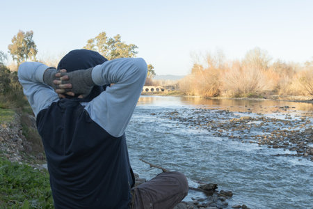 A person is standing by the river, hands placed behind their head, admiring the scenic view. The setting includes water, sky, plants, trees, grass, and a photographer with a reflex cameraの写真素材