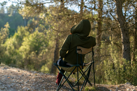 Person relaxes in forest on folding chair, wearing hooded jacket, surrounded by lush green treesの写真素材