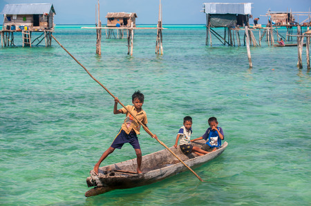 Sea Gypsy Kids on their sampan with their house on stilts in the backgroundのeditorial素材