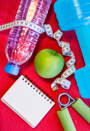 Lay Flat - Dumbbell, Measuring Tape, Hand Grip, Mineral Water, Fresh Apple & Blank Notebook for Motivational Quotes/Words on red background - Fitness Conceptの写真素材
