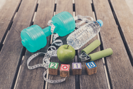 2018 Fitness Concept - Dumbbell, Measuring Tape, Mineral Water, Fresh Apple, Hand Grip & "2018" Wooden Blocks on a wooden table background. Motivation, goals and new year resolution conceptの写真素材