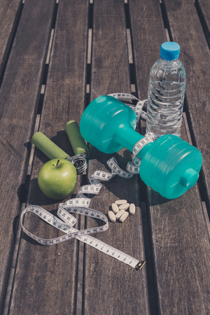 Fitness Concept - Dumbbell, Measuring Tape, Mineral Water, Fresh Apple, Hand Grip & vitamins/supplement tablets on a wooden table background. Motivation, goals and new year resolution conceptの写真素材