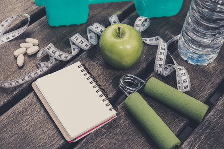 Fitness Concept - Dumbbell, Measuring Tape, Mineral Water, Fresh Apple, Hand Grip, Vitamins Supplement & Blank notebook on a wooden table background. Motivation, goals and new year resolution concept. Shallow depth of field, selective focusの写真素材