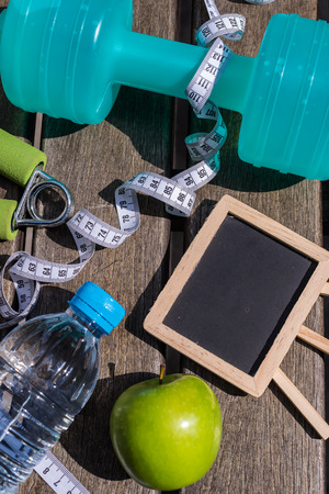 Fitness Concept - Dumbbell, Measuring Tape, Mineral Water, Fresh Apple, Hand Grip & Blank blackboard on a wooden table background. Motivation, goals and new year resolution conceptの写真素材