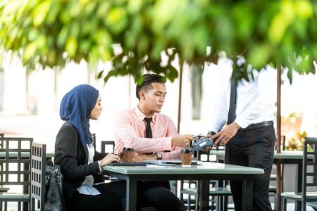Waiter interacting with Muslim customers using bank terminal to process and acquire mobile payment at a coffee shop on a sunny day. Modern cafe start up small business micropayment concept.の写真素材