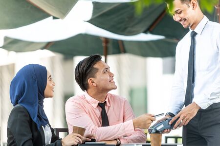 Waiter interacting with Muslim customers using bank terminal to process and acquire mobile payment at a coffee shop on a sunny day. Modern cafe start up small business micropayment concept.の写真素材