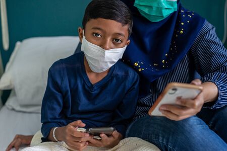 Muslim Mum and Son wearing masks at a hospital, looking at their smartphone. Covid-19 Coronavirus conceptの写真素材