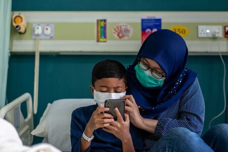 Muslim Mum and Son wearing masks at a hospital, looking at their smartphone. Covid-19 Coronavirus conceptの写真素材