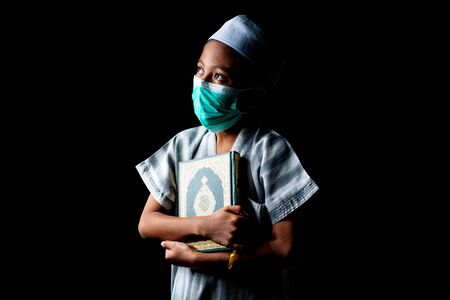 Muslim Boy wearing a surgical mask holding a Quran. Covid-19 coronavirus concept. Copy space for text.の写真素材