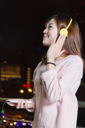 Malaysian female wearing yellow headphones and holding cell phone  with city lights in backgroundの写真素材