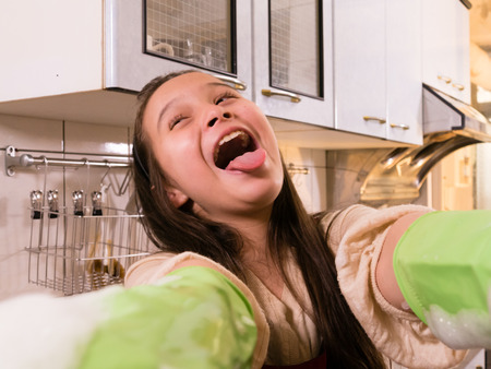 Tween Asian American girl washing dishes, showing foamy gloves with funny expressionの写真素材