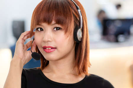 Taiwanese Chinese female wearing a headset with microphone with defocused office or call center in...