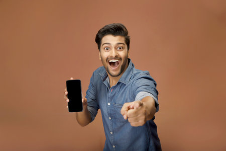 A guy in a shirt is showing his mobile phone screen with one hand and pointing a finger towards the camera, making excited facial expressions.の写真素材