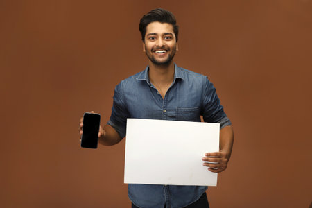 A smart young man is standing holding a blank piece of paper in one hand and a cell phone in the other.の写真素材