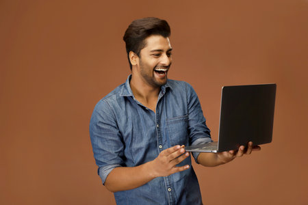 A fashionable guy in casual clothing carrying a laptop in his hands while standing against a brown background.の写真素材