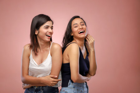 Two beautiful young women laughing and looking at each other on pink backgroundの写真素材