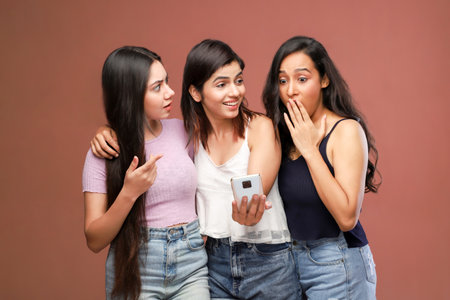 Three young women looking at mobile phone and gossiping on brown backgroundの写真素材