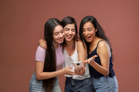 Three happy young women looking at mobile phone while standing against brown backgroundの写真素材