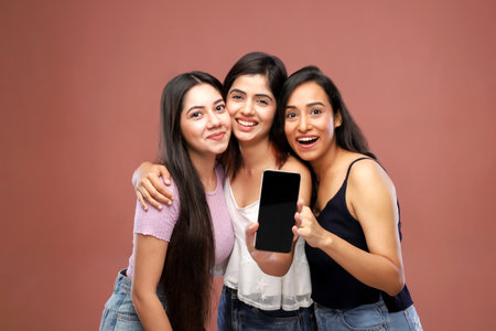 Group of happy young indian women showing smartphone with blank screen, isolated on brown backgroundの写真素材