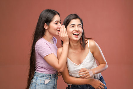 Two young asian women whispering gossip to each other on brown backgroundの写真素材