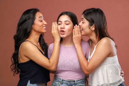 Three young women whispering gossip in each other's ear while standing against brown backgroundの写真素材