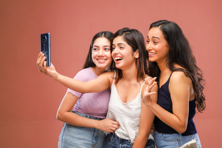 Three young women taking a selfie with a mobile phone isolated on a brown backgroundの写真素材