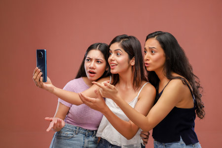 Three young women taking a selfie with a mobile phone. Isolated on brown background.の写真素材