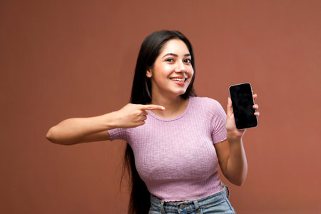 Portrait of a happy young woman pointing at mobile phone against brown backgroundの写真素材