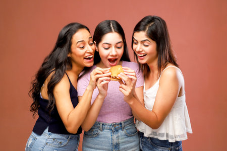Three beautiful young women eating hamburger and laughing on a brown backgroundの写真素材