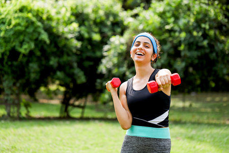 Portrait of smiling young woman lifting dumbbells in the parkの写真素材