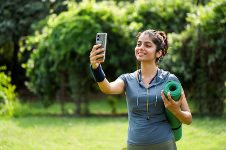 Smiling young woman taking selfie with yoga mat and smartphone in parkの写真素材