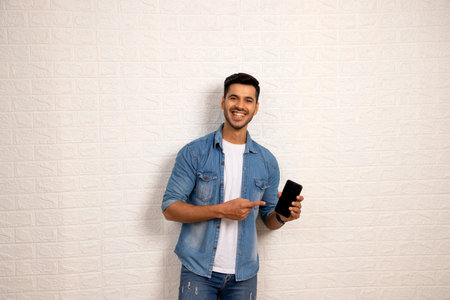 Young man using mobile phone and smiling while standing against white wallの写真素材