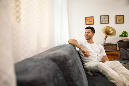 Young man sitting on sofa and drinking coffee in living room at homeの写真素材