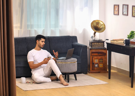 Young man sitting on the floor with laptop and talking on the phoneの写真素材