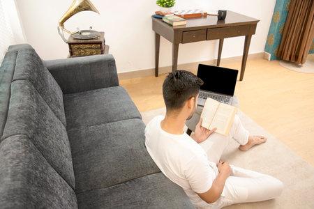 Young man sitting on the floor with a laptop and reading a bookの写真素材