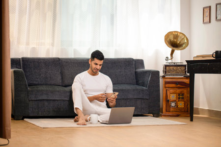 indian man using smartphone while sitting on floor with laptop at homeの写真素材