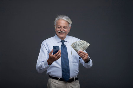 Portrait of mature businessman holding money and using mobile phone on grey backgroundの写真素材