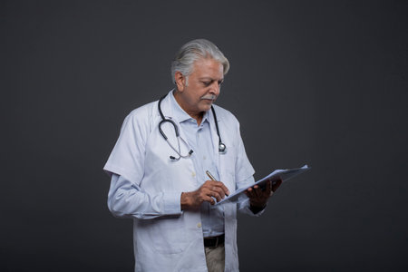 Portrait of senior doctor writing on clipboard while standing against grey backgroundの写真素材