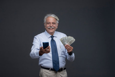 Smiling mature businessman holding money and using mobile phone while standing against grey backgroundの写真素材