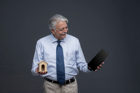 Mature Asian businessman holding a miniature house and a tablet computer on grey backgroundの写真素材