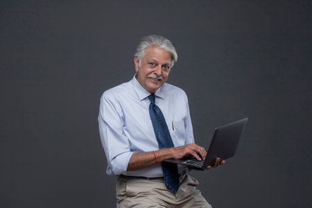 Portrait of happy senior businessman using laptop computer while standing against grey backgroundの写真素材