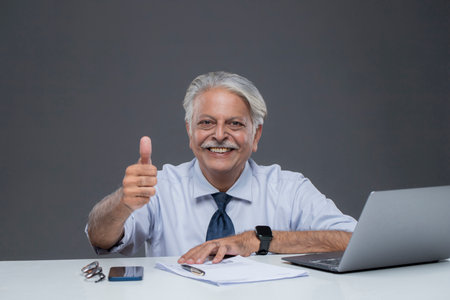 Smiling senior businessman sitting at his desk and showing thumbs up signの写真素材