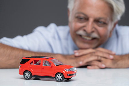 Smiling senior man sitting at table and looking at toy car on grey backgroundの写真素材