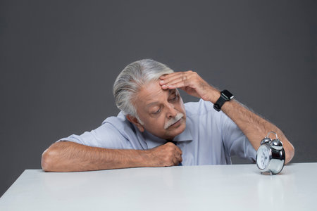 Tired senior man sitting at table with alarm clock on grey backgroundの写真素材