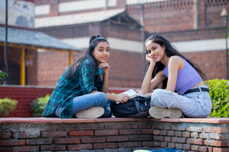 Two female friends sitting on brick wall and looking at camera in collegeの写真素材