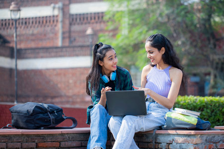 Two young female students sitting on a bench with a laptop and talking in a parkの写真素材