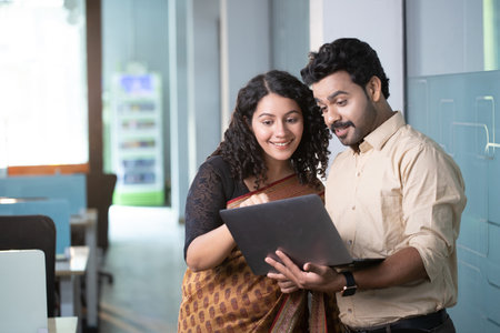 Indian couple using laptop computer in office. They are smiling and looking at cameraの写真素材